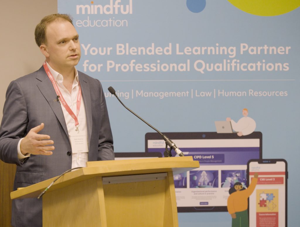 Mindful Education's Managing Director standing in front of a podium, delivering a breakout session about blended learning apprenticeships to delegates at the AoC Annual Conference 2022.