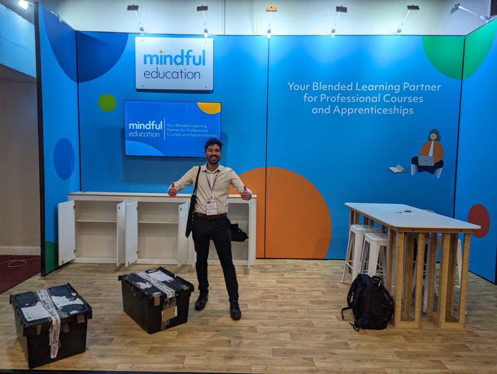 A Mindful Education Employee standing, sticking his thumbs up in front of a Mindful Education branded stand that shows a bag and two moving crates.