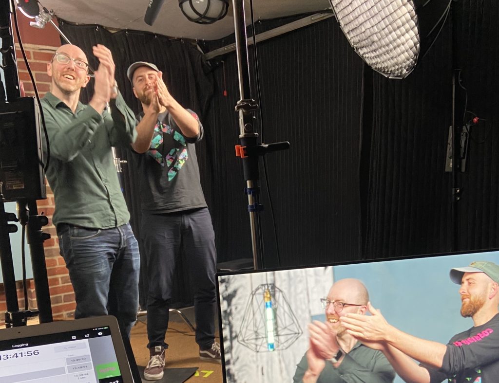 Two Mindful Education employees in a studio with filming equipment, clapping towards the Production team behind the camera.