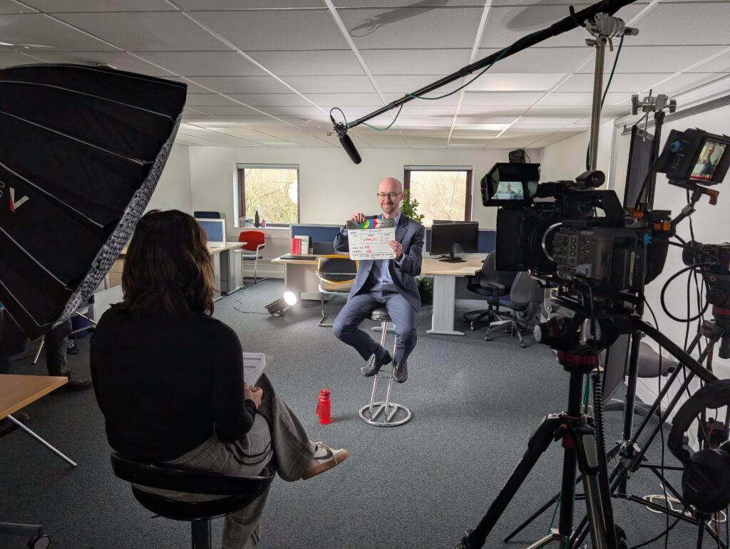 The CEO of a training provider holding a clapperboard in front of filming equipment.